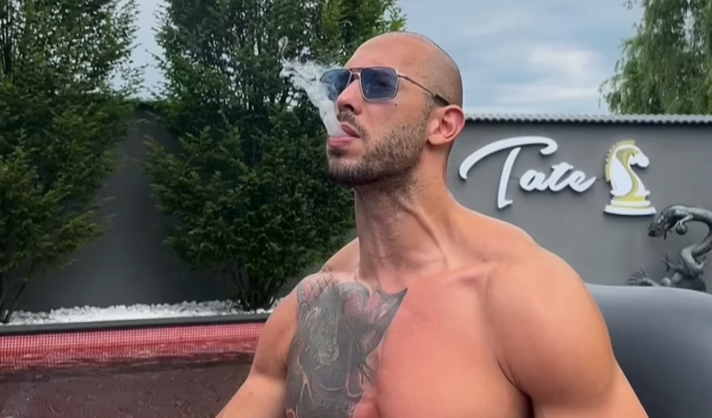 Andrew tate with a bare torso and sunglasses standing outside and smoking